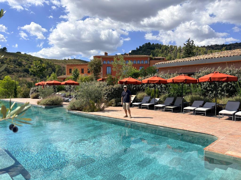Un hombre de pie junto a una piscina en un entorno natural, rodeado de sombrillas naranjas y sillones, con un edificio de estilo rústico al fondo y un paisaje montañoso.