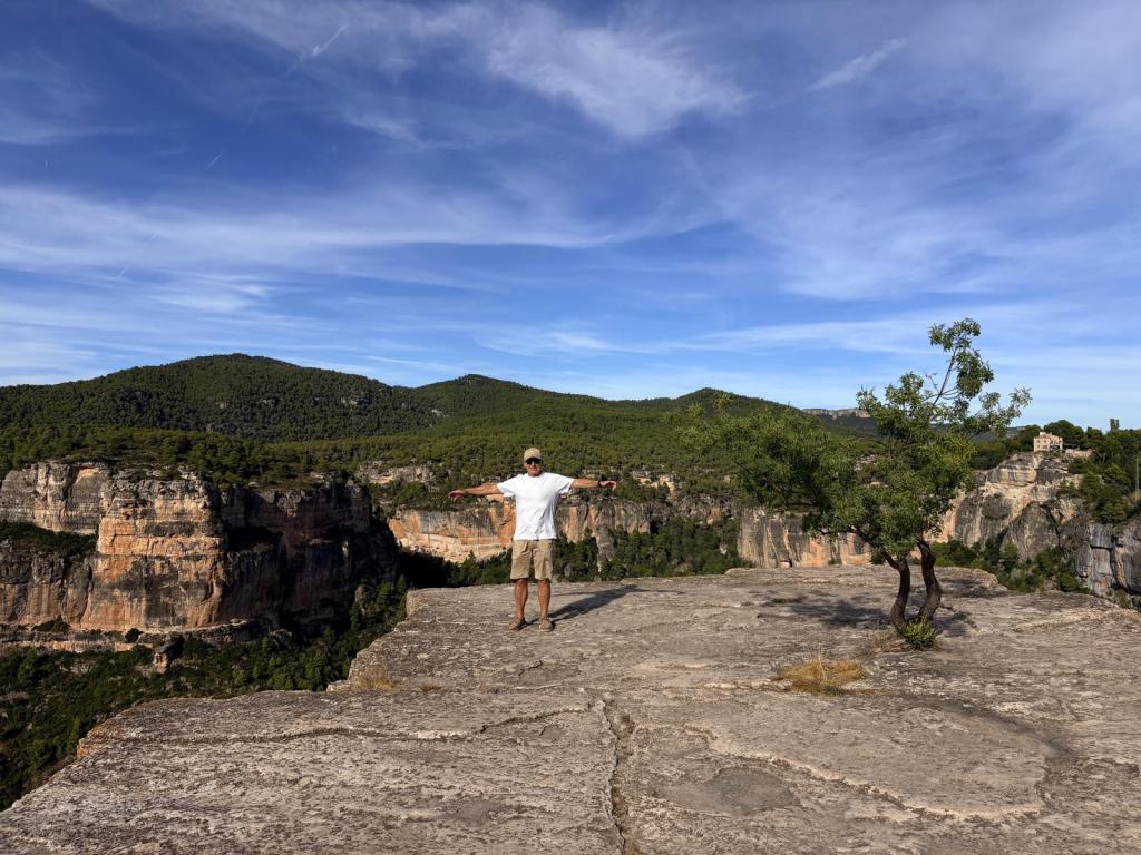 Hombre de pie en un acantilado, con los brazos extendidos, rodeado de un paisaje montañoso y un cielo azul.