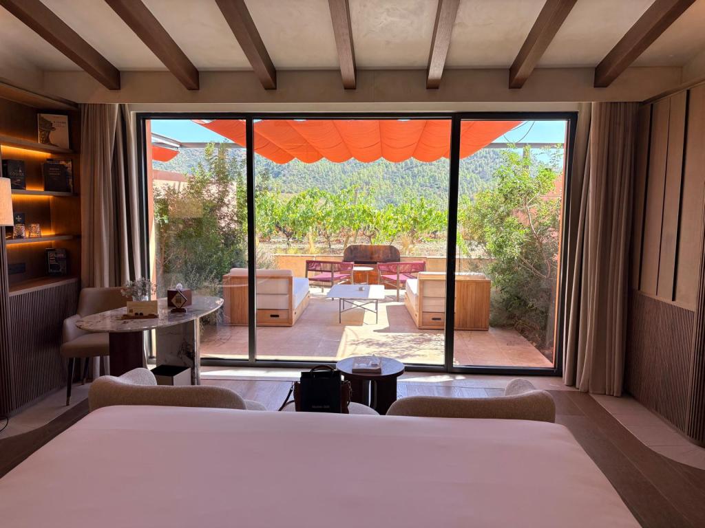 Vista de una habitación luminosa con cama, mesa y sillas, y grandes ventanales que dan a un patio con muebles exteriores y vegetación.