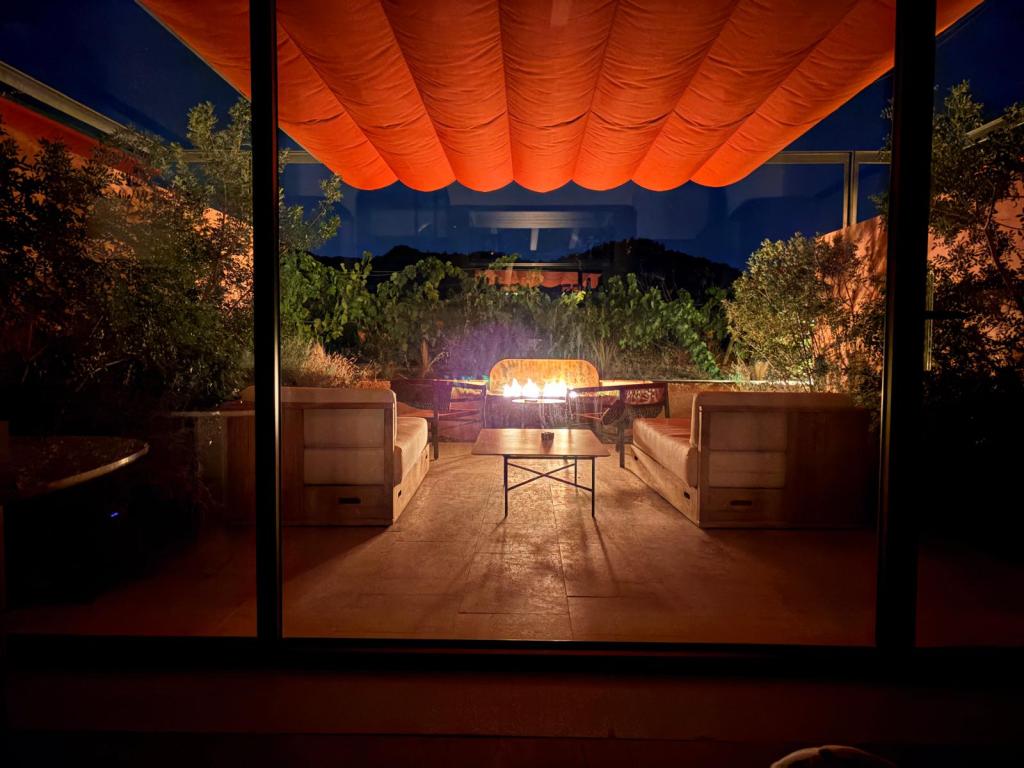 Vista nocturna de una terraza iluminada con un sofá y una mesa de fuego, rodeada de vegetación y un toldo naranja.