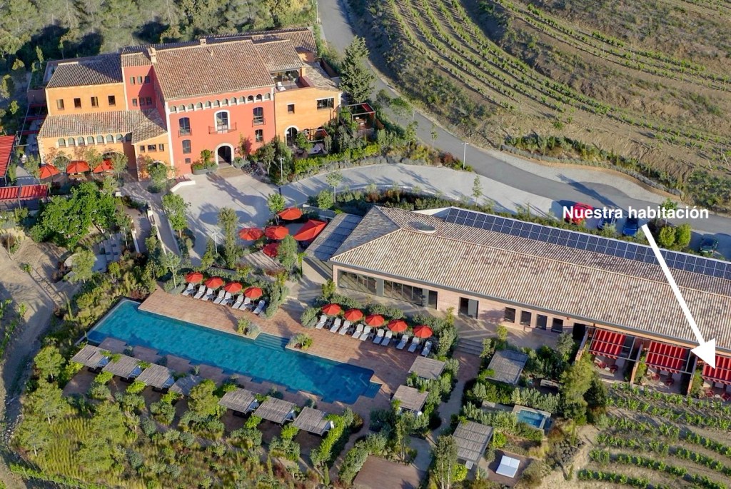 Vista aérea de un complejo hotelero con una piscina, jardines y terrazas. Se destacan edificios de colores cálidos y una señalización que indica 'Nuestra habitación'.