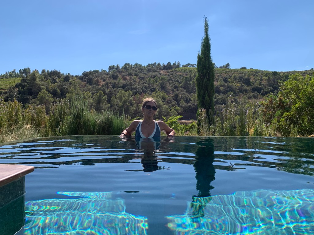 Una mujer relajándose en una piscina con borde infinito, rodeada de vegetación y colinas bajo un cielo azul.