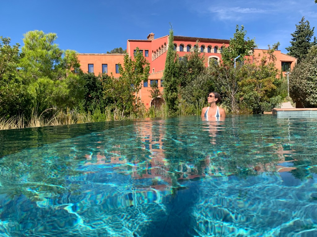 Una mujer relajándose en una piscina de agua clara, con un entorno de vegetación y una casa de color naranja al fondo bajo un cielo azul.